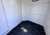 Interior of a small white trailer cargo area with black hooks along the left wall and a rectangular window on the right, dark carpeted floor.