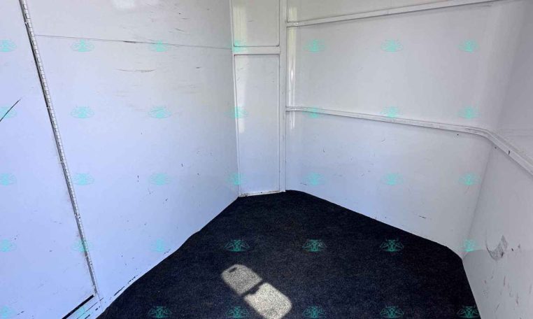 Interior of a small white trailer cargo area with black hooks along the left wall and a rectangular window on the right, dark carpeted floor.