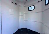Interior of a white horse trailer with a row of hooks on the left wall, a small window on the right, and a ceiling skylight; dark carpeted floor.