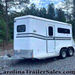 White gooseneck cargo trailer with black stripe accents parked on gravel, surrounded by trees and a chain-link fence in a wooded lot.