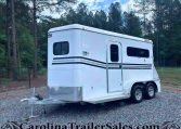 White gooseneck cargo trailer with black stripe accents parked on gravel, surrounded by trees and a chain-link fence in a wooded lot.