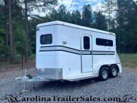 White gooseneck cargo trailer with black stripe accents parked on gravel, surrounded by trees and a chain-link fence in a wooded lot.