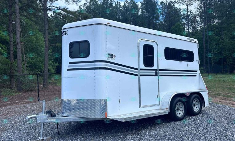 White gooseneck cargo trailer with black stripe accents parked on gravel, surrounded by trees and a chain-link fence in a wooded lot.