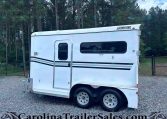 Side view of a white travel trailer on a gravel lot with trees in the background, showing two windows and a door; appears unhooked near a fence.