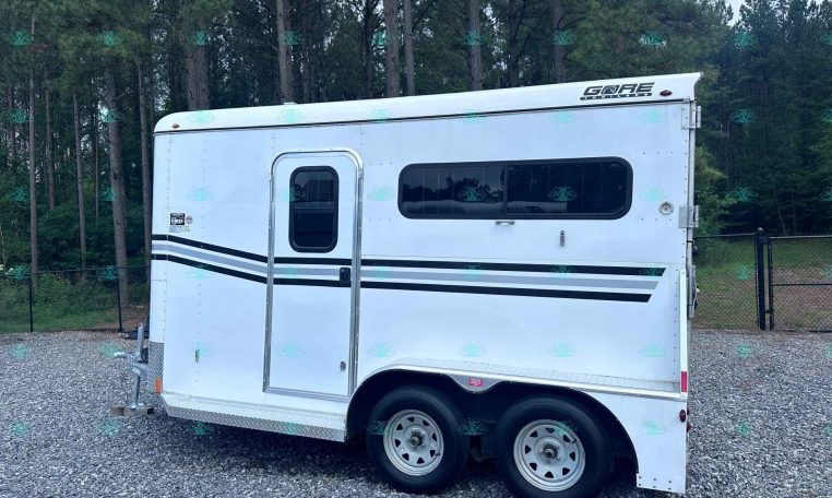 Side view of a white travel trailer on a gravel lot with trees in the background, showing two windows and a door; appears unhooked near a fence.