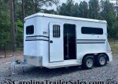 White horse trailer on a gravel lot with the side door open, showing a barred window and two axles; trees form a forest behind it.