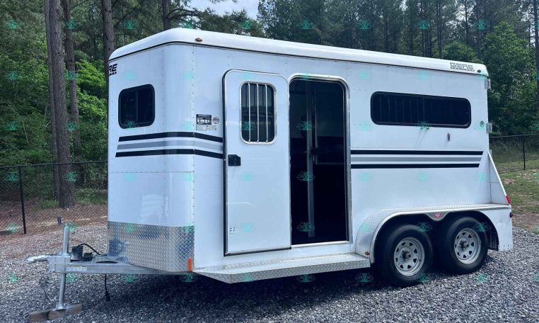 White horse trailer on a gravel lot with the side door open, showing a barred window and two axles; trees form a forest behind it.
