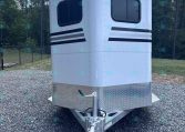Front view of a white enclosed trailer with two windows and black stripe decals, parked on gravel. Maintains hitch and diamond-plate base visible at the bottom.