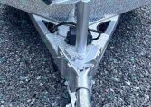 Front of a utility trailer tongue with a manual crank jack, safety chains, and a coupler resting on a gravel surface behind a diamond-plate box railings area.
