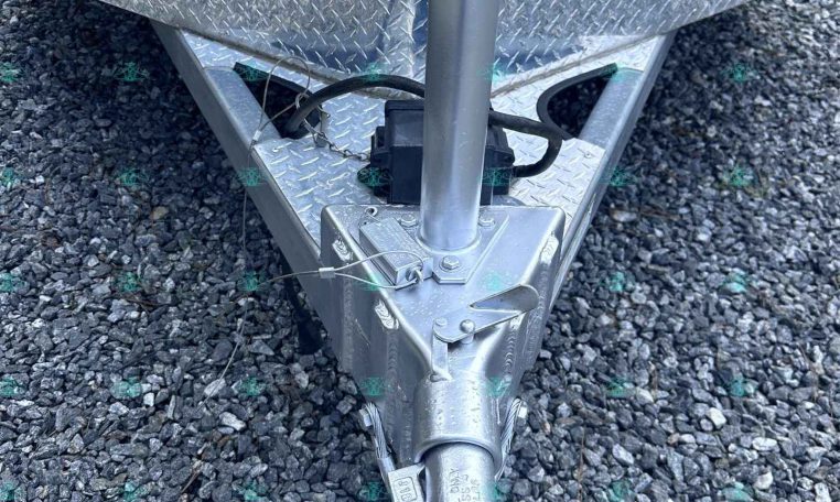 Front of a utility trailer tongue with a manual crank jack, safety chains, and a coupler resting on a gravel surface behind a diamond-plate box railings area.