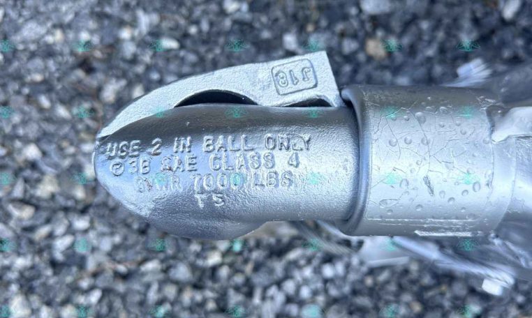 Close-up of a metal trailer hitch coupler with stamped text on a gravel surface, showing specifications and a worn, damp finish.