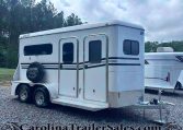 White two-axle bumper-pull horse trailer with multiple windows and a spare tire mounted on the back, on a gravel lot with trees in the background.