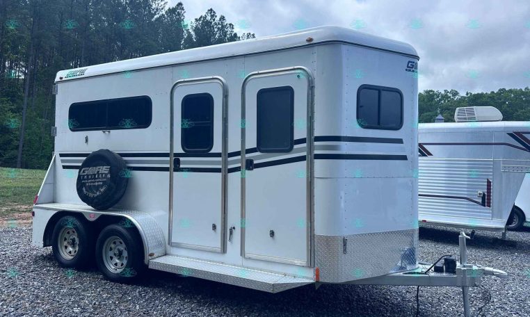 White two-axle bumper-pull horse trailer with multiple windows and a spare tire mounted on the back, on a gravel lot with trees in the background.