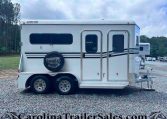 White gooseneck horse trailer with spare tire on a gravel lot, parked at a trailer sales yard with trees in the background.