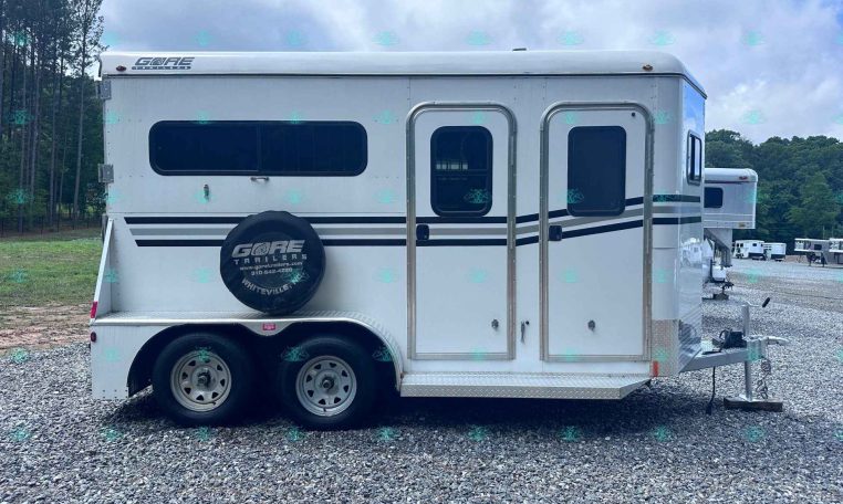White gooseneck horse trailer with spare tire on a gravel lot, parked at a trailer sales yard with trees in the background.