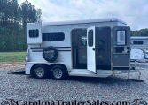 White horse trailer with open side door on a gravel lot, forested background and a spare tire cover on the back tire.