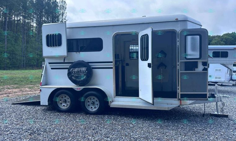 White horse trailer with open side door on a gravel lot, forested background and a spare tire cover on the back tire.