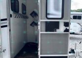 Open travel trailer door showing metal entry steps and interior hallway, with windows along the side and other trailers visible in a gravel lot