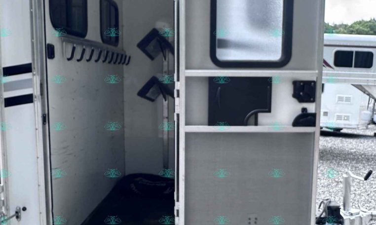 Open travel trailer door showing metal entry steps and interior hallway, with windows along the side and other trailers visible in a gravel lot