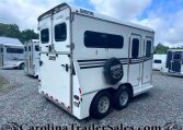 Rear and side view of a white enclosed travel trailer with windows and a spare tire cover in a gravel lot; other trailers are visible in the background.