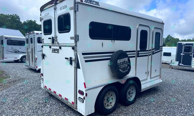 Rear and side view of a white enclosed travel trailer with windows and a spare tire cover in a gravel lot; other trailers are visible in the background.