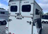 White horse trailer rear with double doors, two small rear windows, exterior latches, and a spare tire mounted on the side in a gravel lot.