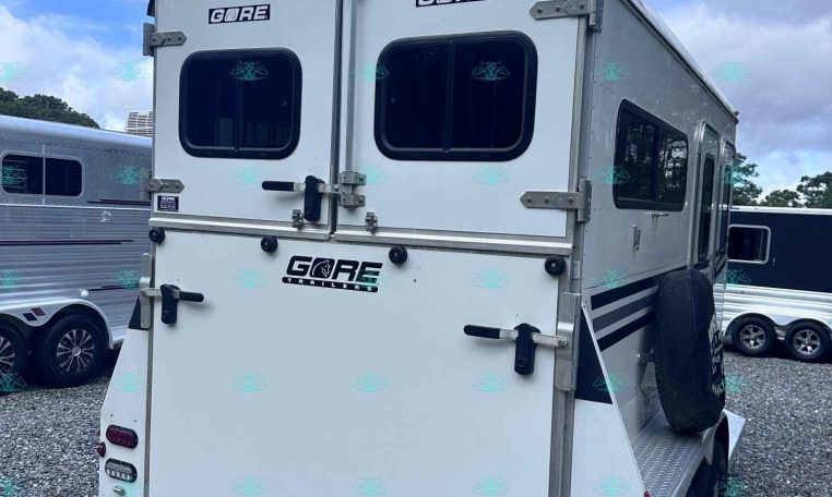 White horse trailer rear with double doors, two small rear windows, exterior latches, and a spare tire mounted on the side in a gravel lot.