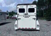 Rear view of a white horse trailer in a gravel lot with two rear doors and small windows, with other trailers and trees in the background.