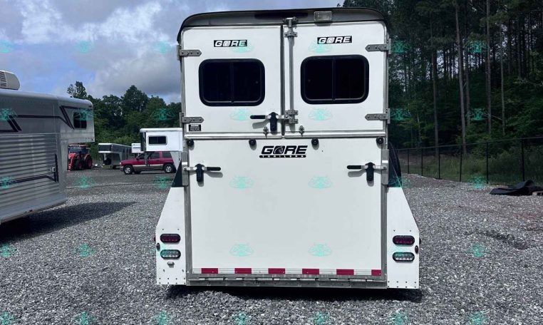 Rear view of a white horse trailer in a gravel lot with two rear doors and small windows, with other trailers and trees in the background.
