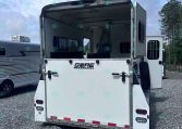 White horse trailer with rear doors open, showing interior stalls and dividers, parked on a gravel lot beside another trailer under a blue sky