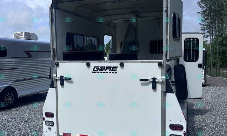 White horse trailer with rear doors open, showing interior stalls and dividers, parked on a gravel lot beside another trailer under a blue sky