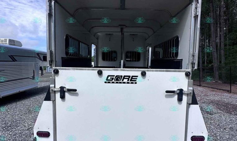 Open rear of a white horse trailer with interior stalls visible, parked on a gravel lot under a blue sky with trees nearby.