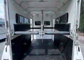 Empty horse trailer interior with dividing partitions, padded walls, and side windows; rear ramp lowered on a gravel surface.