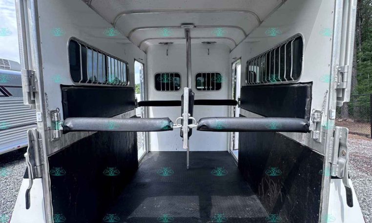 Empty horse trailer interior with dividing partitions, padded walls, and side windows; rear ramp lowered on a gravel surface.