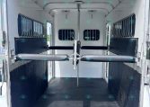 Interior of a white horse trailer with padded walls, metal partitions, and barred windows; empty with a diamond-plate rubber floor.