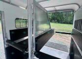 Interior of a metal horse trailer with a partition and rubber-matted floor, viewed from the doorway toward outside grass and trees outside the window.