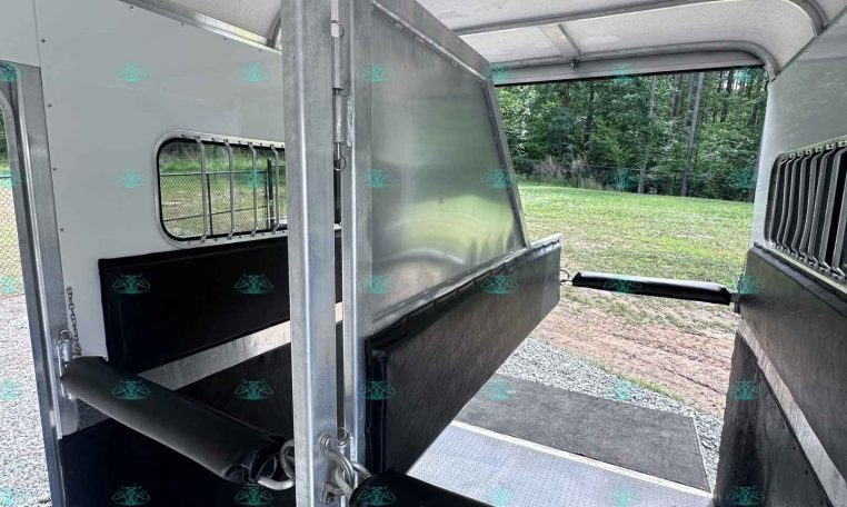 Interior of a metal horse trailer with a partition and rubber-matted floor, viewed from the doorway toward outside grass and trees outside the window.