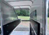 Interior of a horse trailer with metal dividers and cushioned walls, looking toward exterior field through the rear opening.
