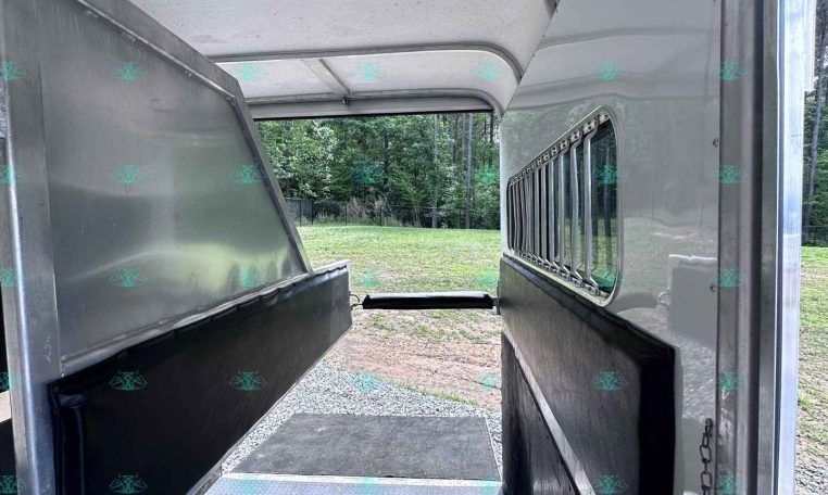 Interior of a horse trailer with metal dividers and cushioned walls, looking toward exterior field through the rear opening.