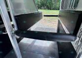 Interior of a horse trailer showing a padded divider on the left, metal walls, and a diamond-plate floor with the rear doors open to a grassy, tree-filled outdoor scene.