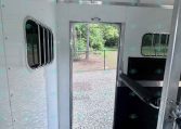 Interior of aluminum trailer with open back door, showing gravel ground, chain-link fence, and trees outside.
