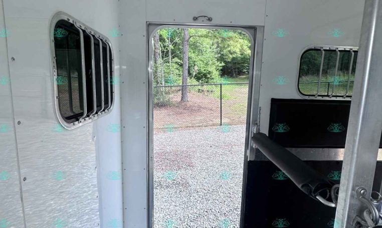 Interior of aluminum trailer with open back door, showing gravel ground, chain-link fence, and trees outside.