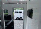 Interior of a silver horse trailer looking out the open door to a gravel lot with another white trailer outside.