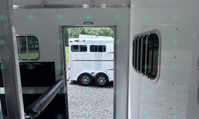 Interior of a silver horse trailer looking out the open door to a gravel lot with another white trailer outside.