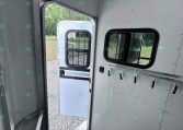 Interior of a light-gray enclosed trailer with the side door open, showing wall hooks, a windowed wall, and a diamond-plate floor; outside gravel and greenery visible.
