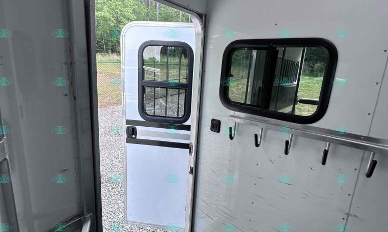 Interior of a light-gray enclosed trailer with the side door open, showing wall hooks, a windowed wall, and a diamond-plate floor; outside gravel and greenery visible.