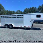 Silver aluminum horse trailer with multiple side windows parked on a gravel lot, backed by pine trees and a blue building under a clear sky.