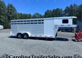 Silver aluminum horse trailer with multiple side windows parked on a gravel lot, backed by pine trees and a blue building under a clear sky.