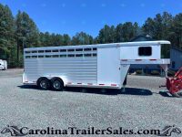 Silver aluminum horse trailer with multiple side windows parked on a gravel lot, backed by pine trees and a blue building under a clear sky.