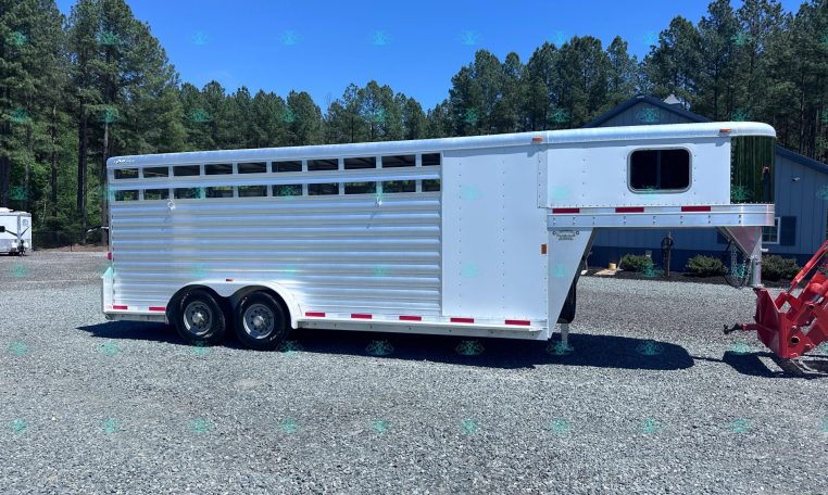 Silver aluminum horse trailer with multiple side windows parked on a gravel lot, backed by pine trees and a blue building under a clear sky.